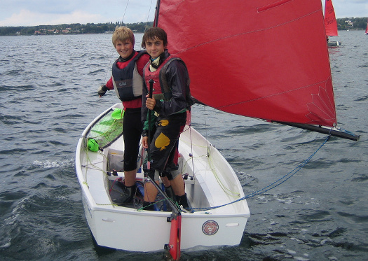 A white Mirror dinghy lying-to with helm & crew looking aft and smiling at the camera
