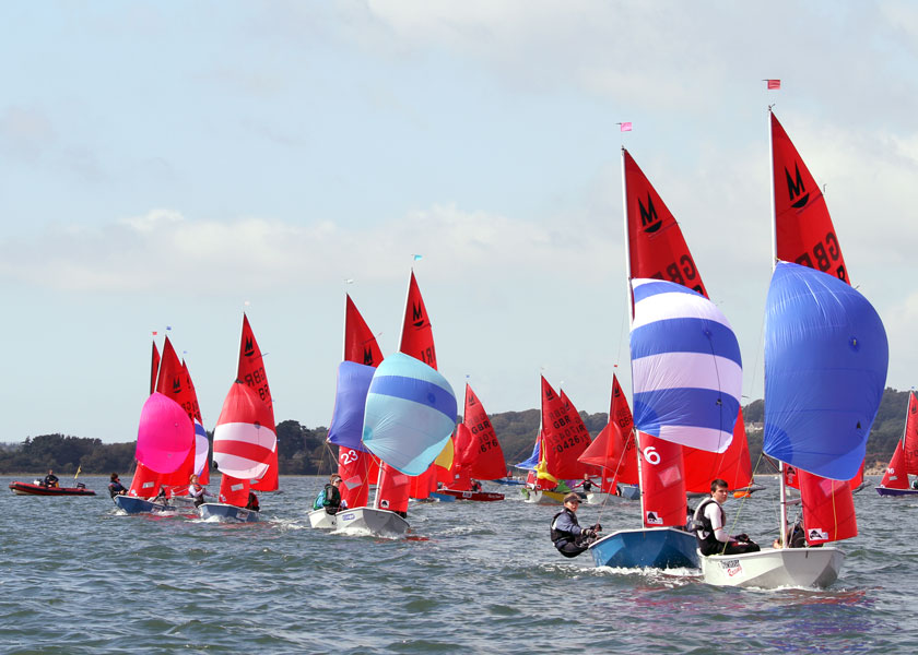 A fleet of Mirror dinghies racing with the windward mark in the background and boats with spinnaker flying sailing towards the camera