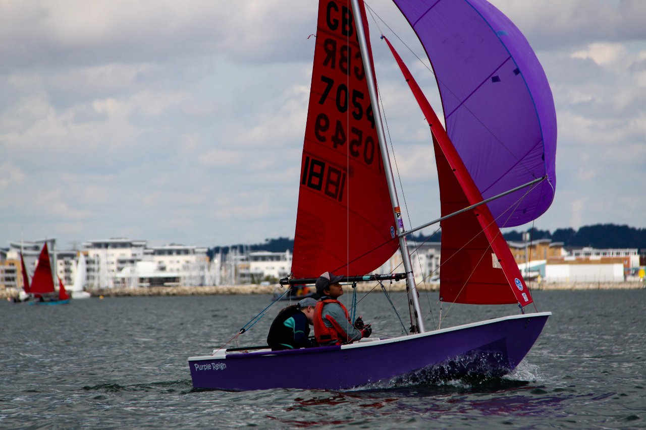 A purple Mirror with purple spinnaker sailing across the picture under a gloomy sky
