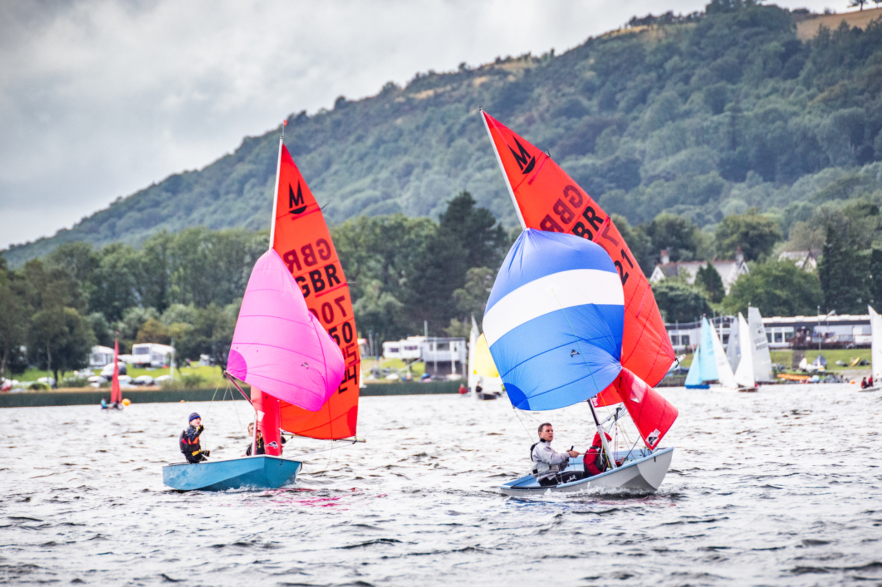 Two Mirror dinghies racing close together with spinnakers set with a sailing club, other boats, in the distance and the hills of the lake district in the background