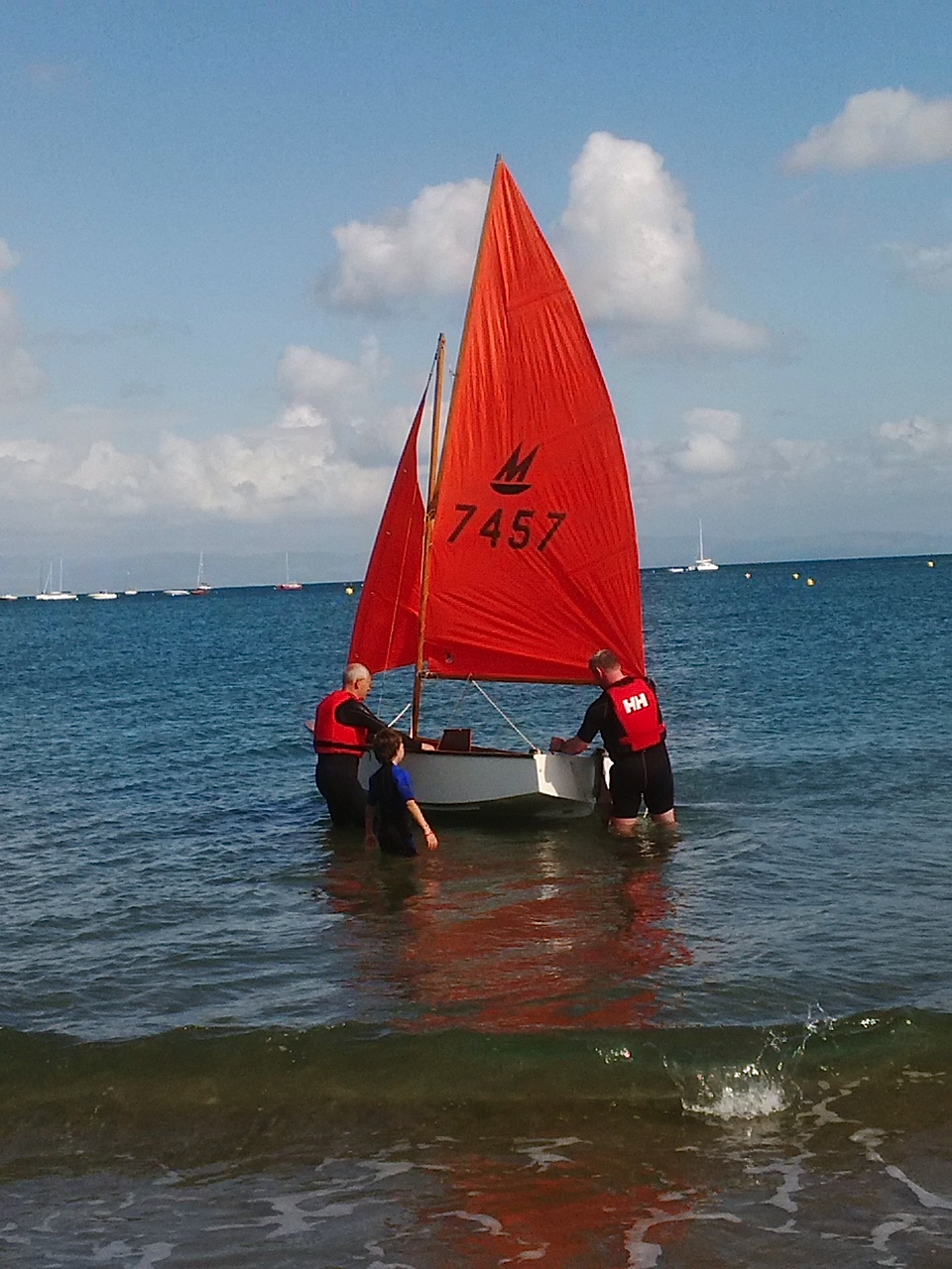 A white wooden Mirror dinghy being held in knee deep water by two gentleman and a child in wetsuits prior to launching