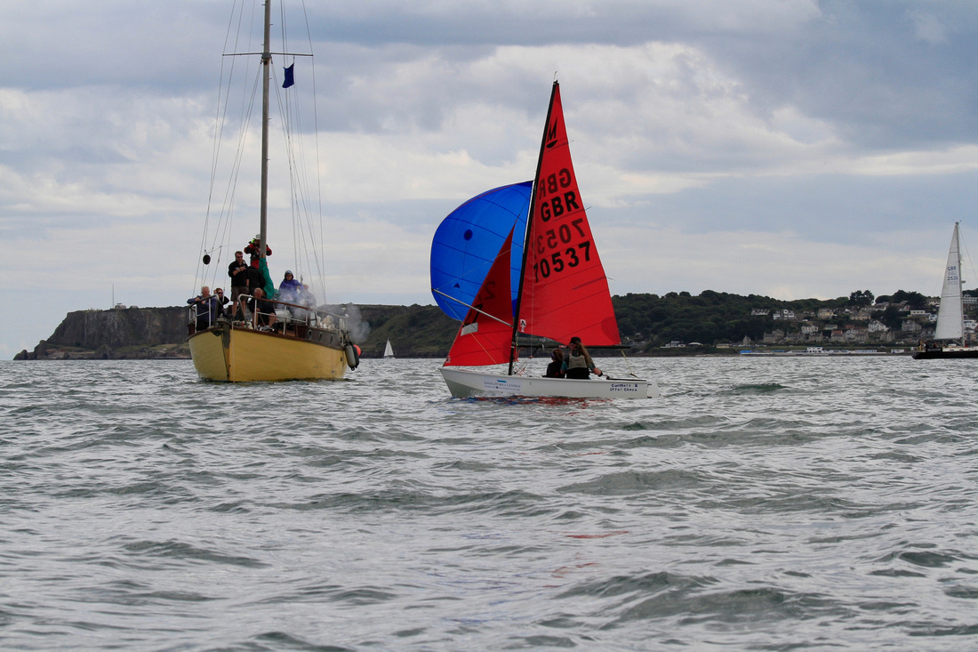 A white GRP Mirror dinghy with spinnaker set crosses the finish line with smoke from the cannon on the Committee boat visible